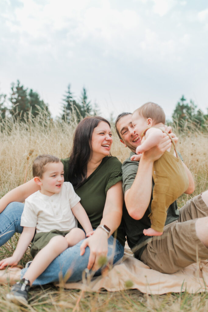 A family of four with two young kids sit snuggled up and smiling at eachother. They backdrop is a beautiful field.