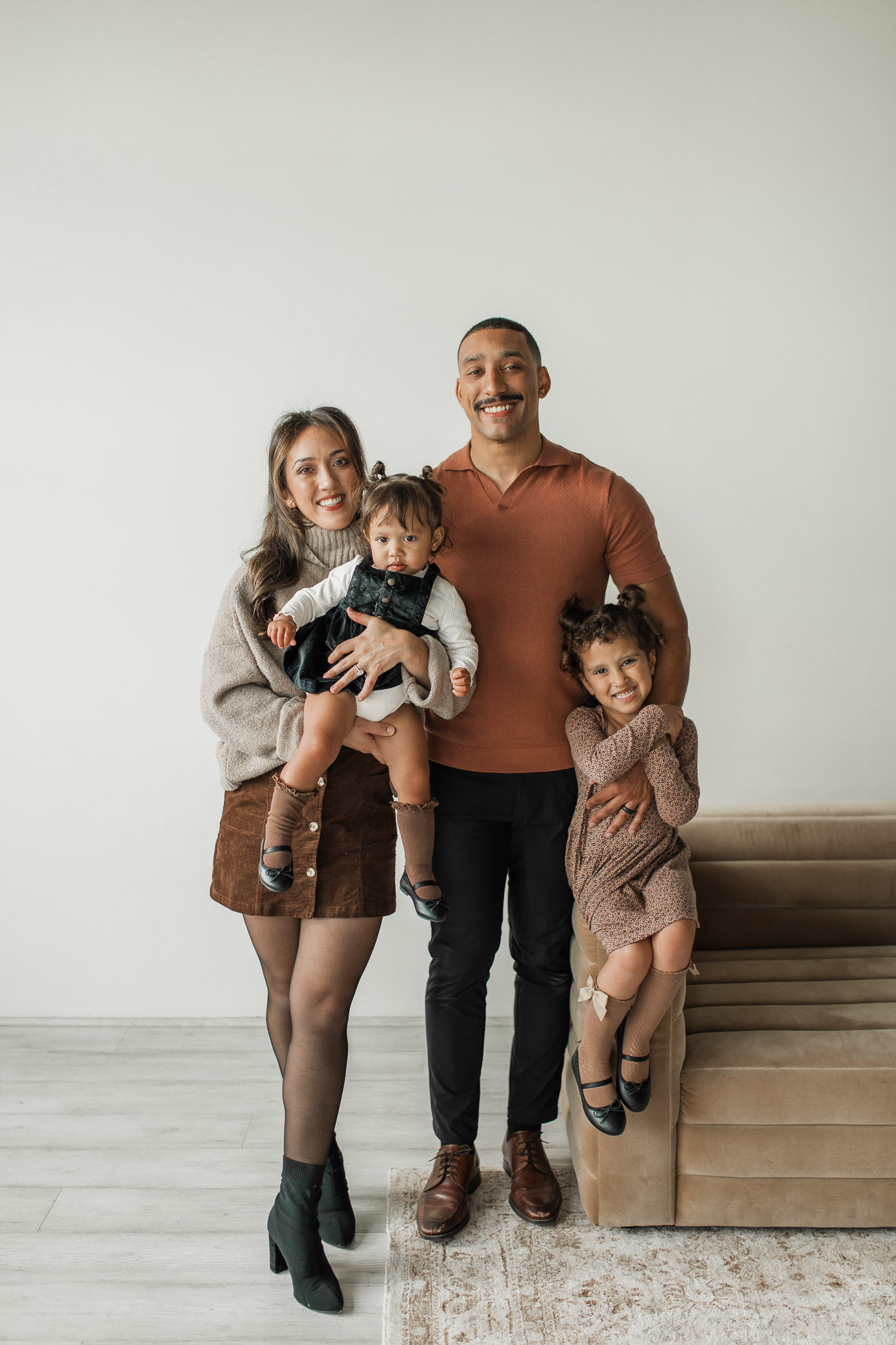 In studio family photo featuring a family or four with two toddler girls wearing nuetral browns and tans. All four people are smiling at the camera.