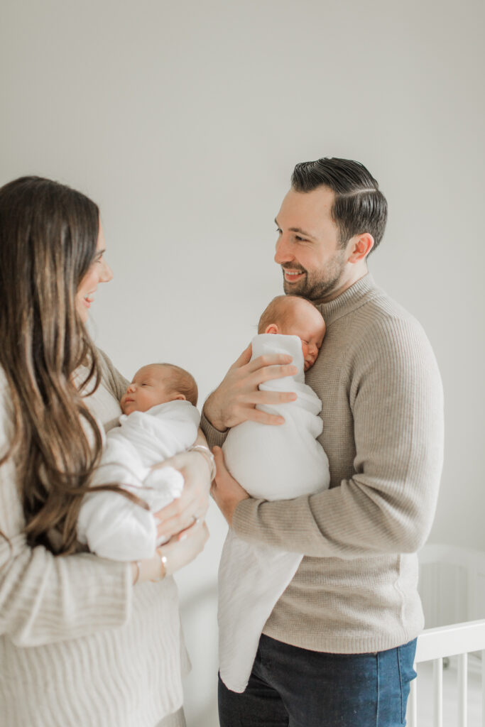 Newborn outfit photoshoot with white swaddles on twin boys and parents wearing neutral sweaters.