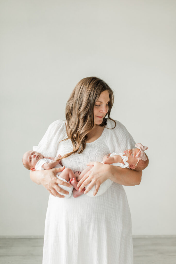 Newborn photos in studio with the mother holding both babies in her arms, looking lovingly at one baby.