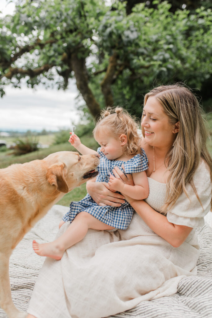 backyard family photo with a mother holding her toddler girl and a dog licking her