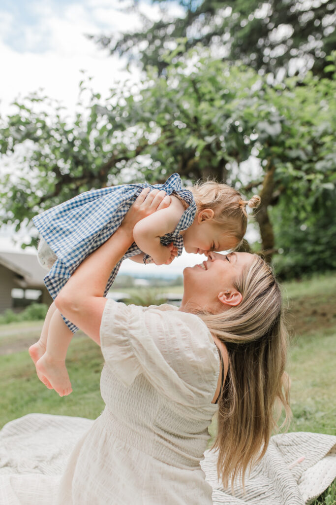a mother holds up her toddler daughter, lovingly touching their noses together for backyard family photos. her long blond hair flows back.