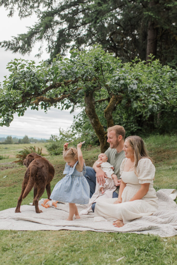 backyard family photo featuring a toddler girl twirling in her dress while her family and pet dog sit on a picnic blanket.