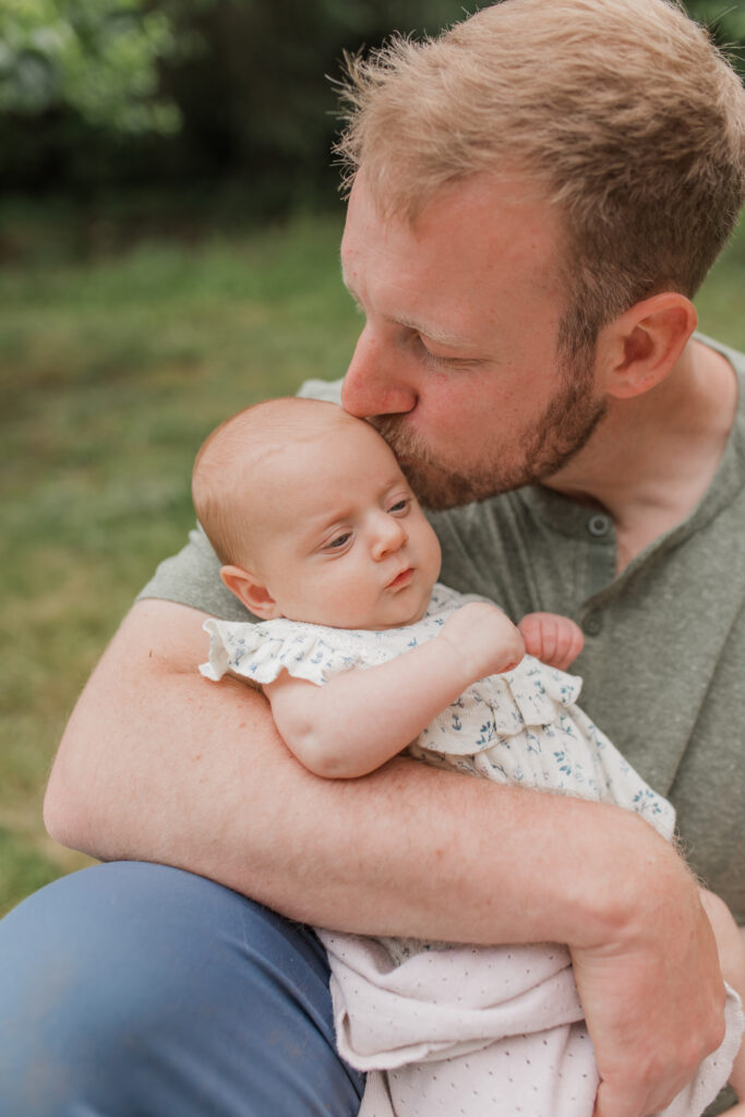 father holds his newborn baby girl and kisses her head in their backyard for backyard family photos.