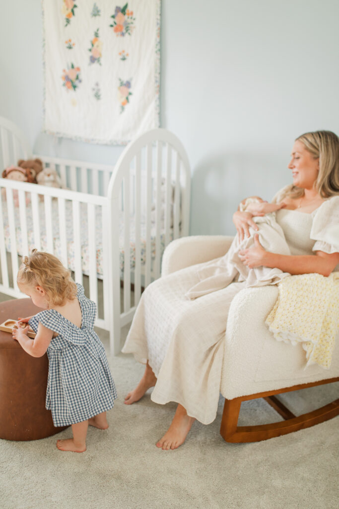 a mom rocks her newborn baby in a rocking chair in the nursery while a toddler girl plays with a wooden puzzle nearby.