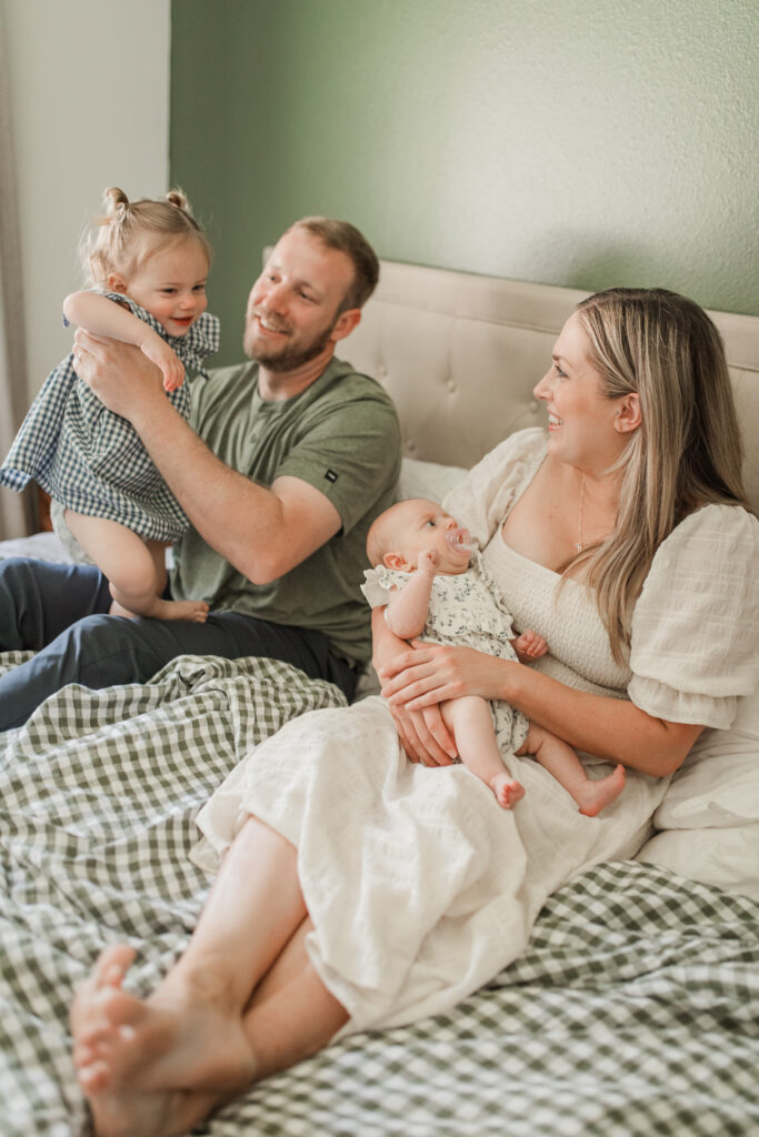 a family snuggles on their king size bed and laughs all together while holding their toddler and baby daughters.