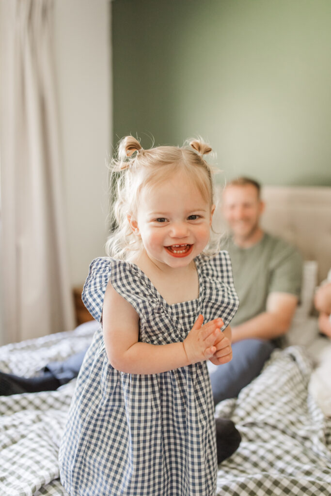 toddler girl jumps and laughs on the bed with her parents behind her.