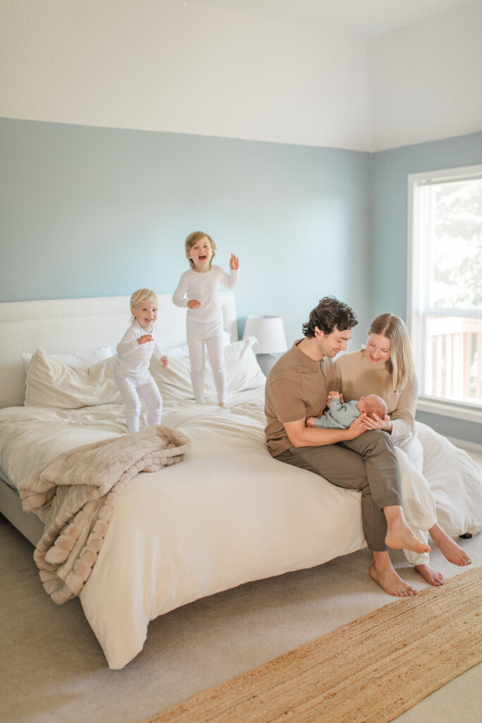 in-home lifestyle newborn photo session with toddlers jumping on the bed and parents sitting, holding their newborn baby
