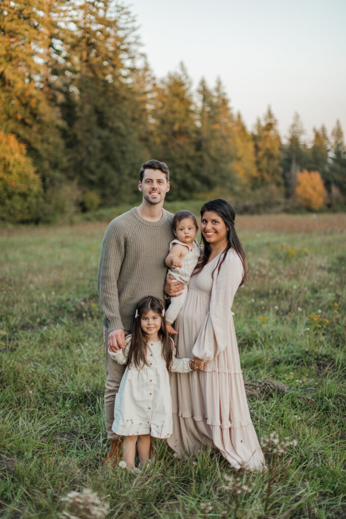 a beautiful family poses in front of a field of trees with changing fall colored leaves. they are wearing long sleeves, corduroy dress and cozy knit sweaters.