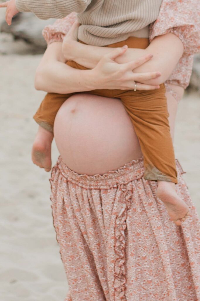 Mom holding toddler hands with baby bump visible on the beach