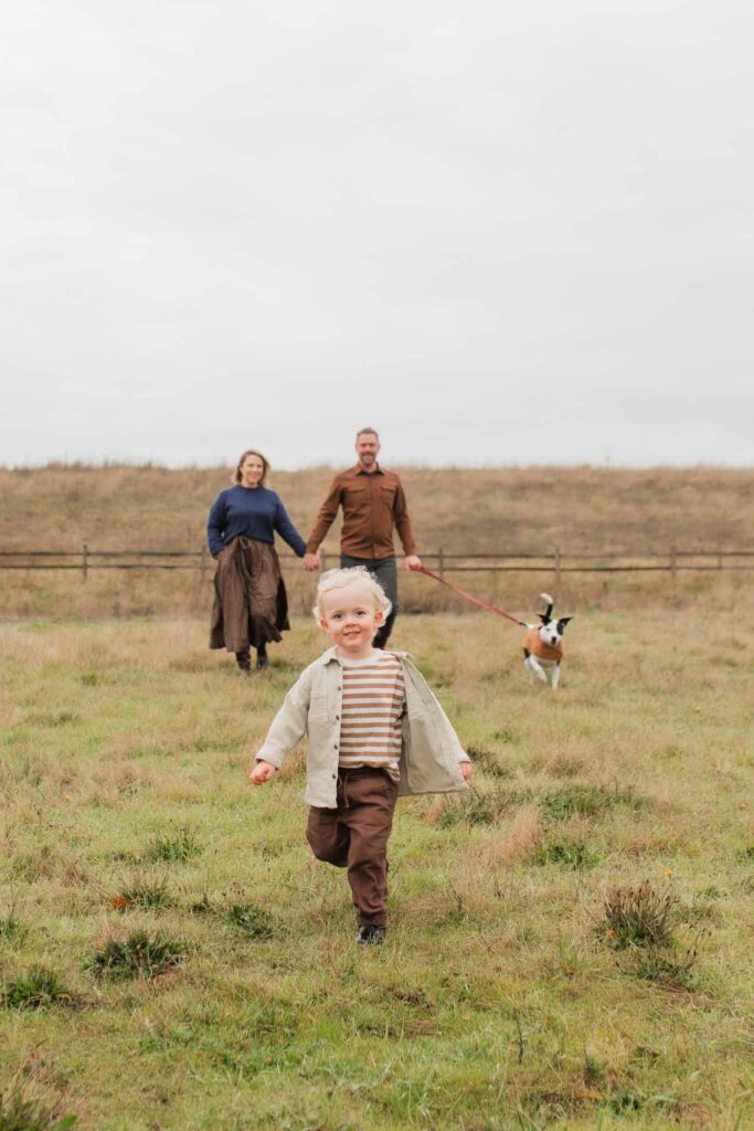 Family of three enjoying fall at Powell Butte, toddler running through a golden field with autumn trees in the background.