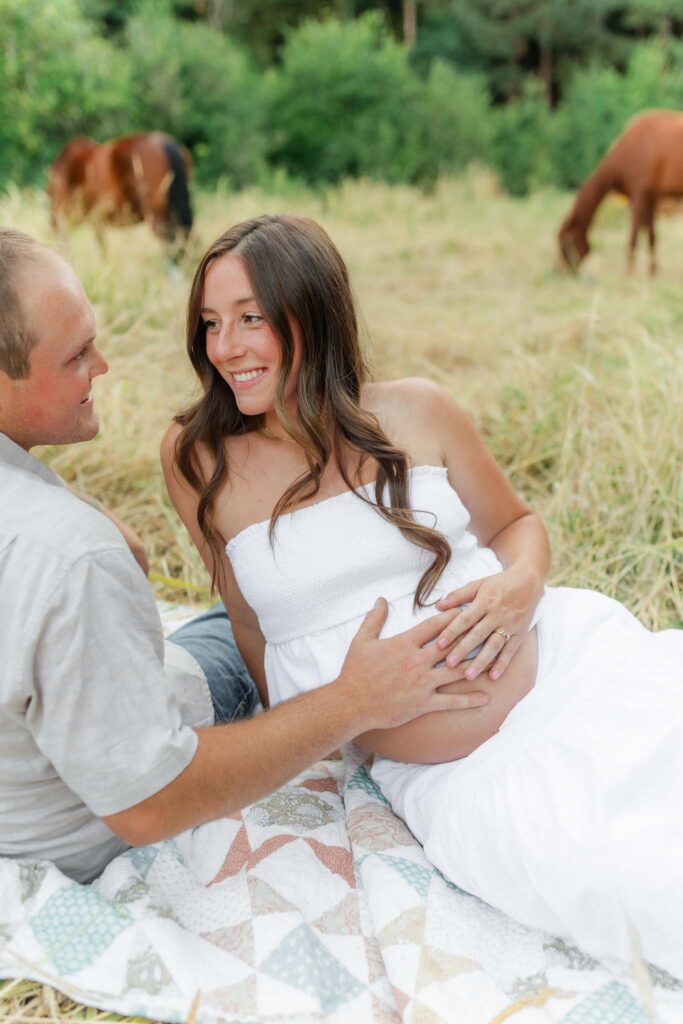 Couple embracing during a golden field maternity session with horses at sunset