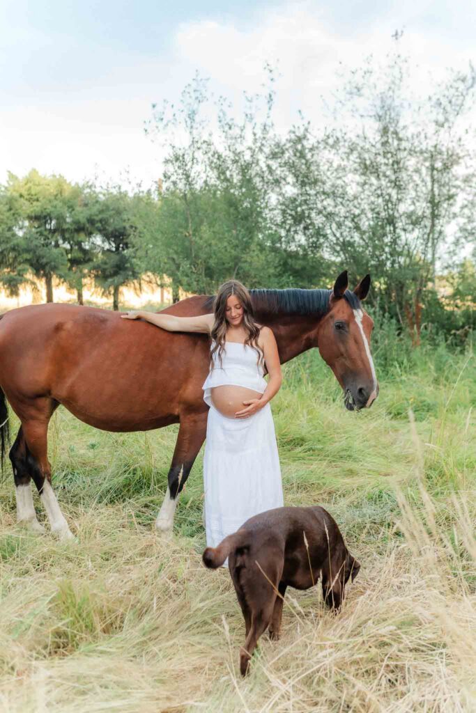 Expecting mother standing beside her horse and dog in a field during a western-inspired maternity photo session
