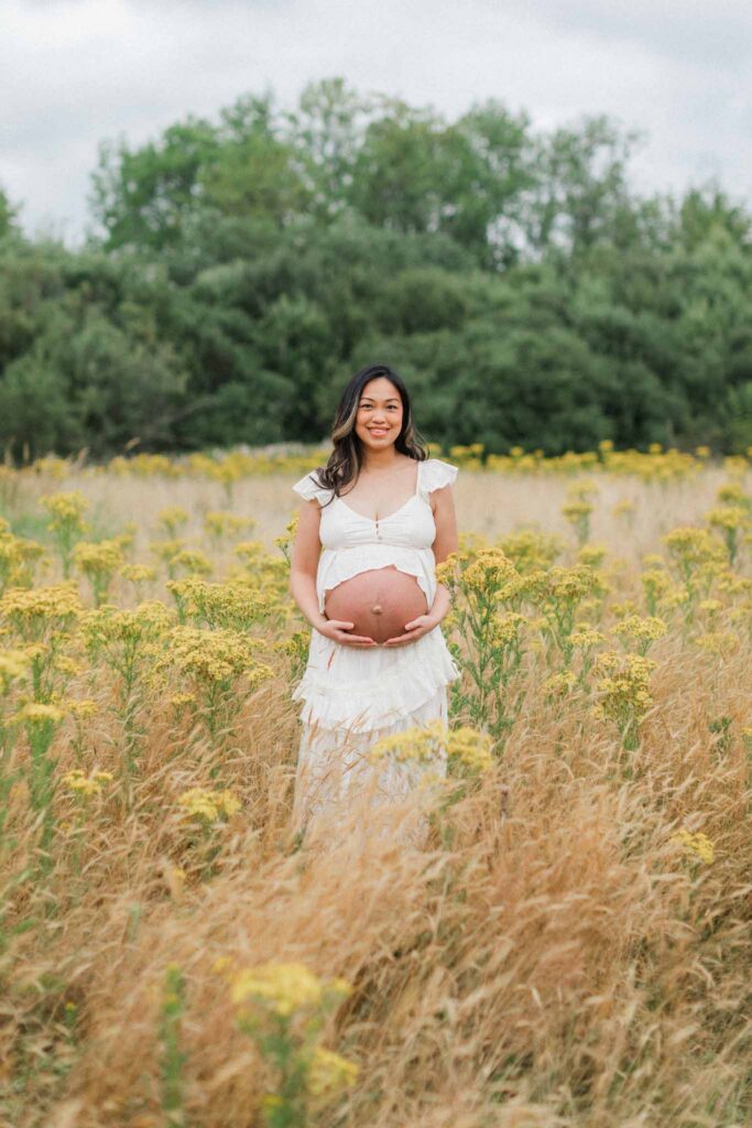 flower field maternity photo with a mom smiling and holding her bump with both arms.